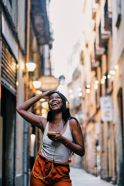 Fröhliche Afroamerikanerin hört Musik mit Handy und Kopfhörer im Freien. - Foto, Bild