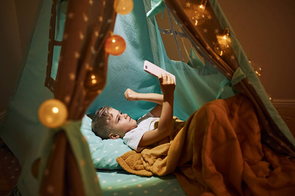 Використовувати телефон. Молодий хлопчик в повсякденному одязі лежить в наметі в вечірній час
. - Фото, зображення