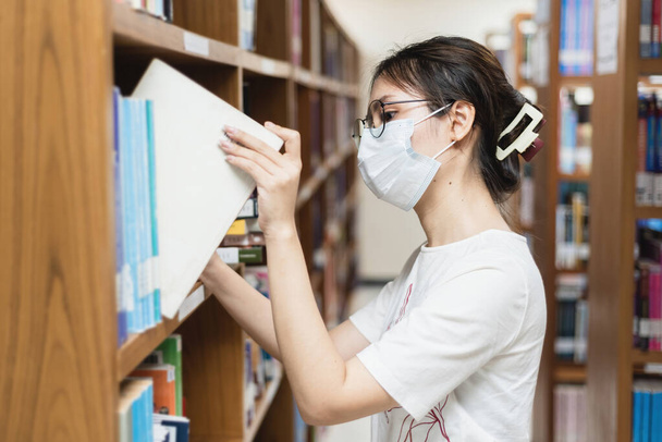 In the library, an Asian woman wearing a hygiene mask is picking up books. - Foto, immagini