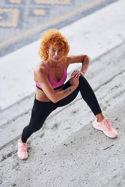 Young european redhead woman in sportive clothes doing squats outdoors. - Foto, imagen