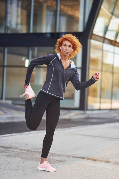 Young european redhead woman in sportive clothes doing exercises outdoors. - Фото, изображение