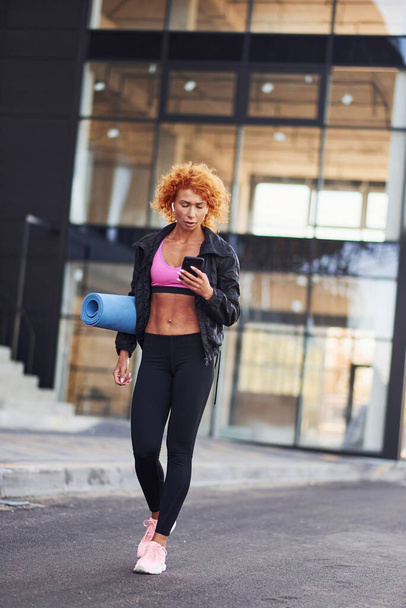 Young european redhead woman in sportive clothes with phone and mat in hands outdoors. - Zdjęcie, obraz
