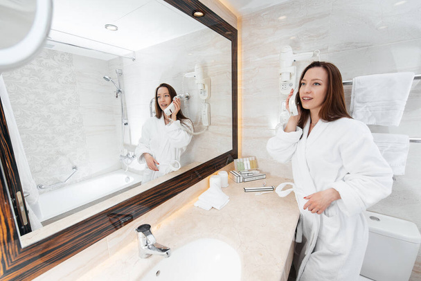 A young beautiful girl in a beautiful white bathroom talking on the phone. A fresh good morning at the hotel. Rest and travel. Hotel recreation, and tourism. - Foto, Bild