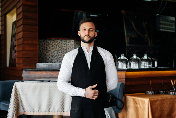 Ein junger Kellner mit Bart zieht sich eine Schürze an und bereitet sich auf einen Arbeitstag in einem feinen Restaurant vor. - Foto, Bild
