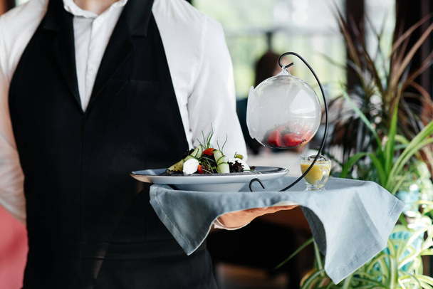 A young waiter in a stylish uniform stands with an exquisite dish on a tray near the table in a beautiful restaurant close-up. Restaurant activity, of the highest level. - Foto, Imagen