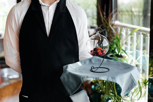 A young waiter in a stylish uniform stands with an exquisite dish on a tray near the table in a beautiful restaurant close-up. Restaurant activity, of the highest level. - Fotó, kép