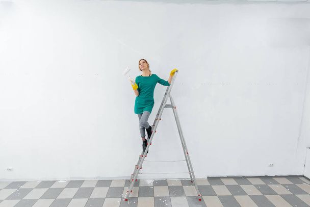 A young girl is engaged in repairs on the stairs and paints a white wall with a roller in a new apartment. Renovation of the interior and a new apartment. Housewarming and a desirable mortgage. - Photo, image