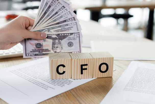 cropped view of woman holding dollars near cubes with cfo lettering on papers on desk - Photo, Image