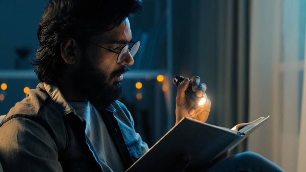 Bearded adult man in glasses at night home living room with flashlight shines on book reads preparing for exams reading bible, faith concept. 30s guy enjoying history in textbook novel at nighttime - Photo, image