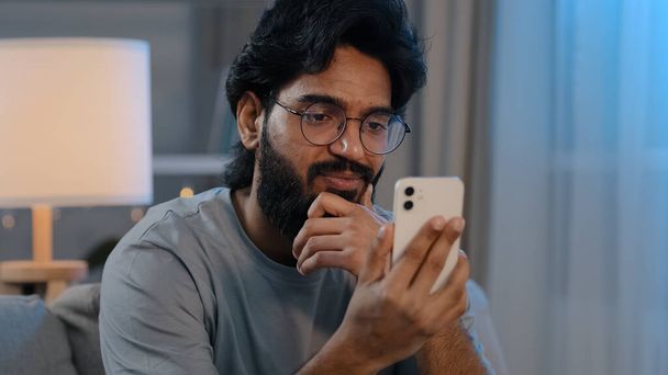 Arabic bearded 30s man in glasses at home with mobile phone talking on video call conference chat distance talking using smartphone for casual virtual communication with friends conversation online - Foto, imagen