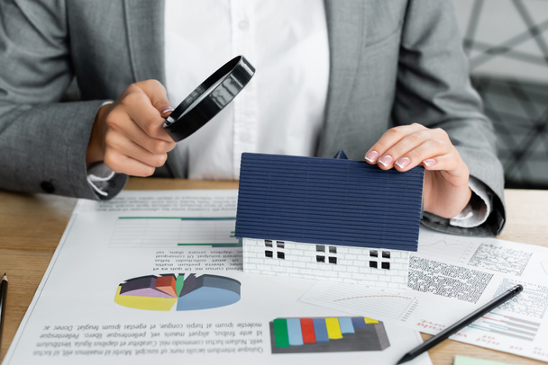 oříznutý pohled na daňového inspektora držícího lupu nad modelem domu v blízkosti grafů na stole - Fotografie, Obrázek