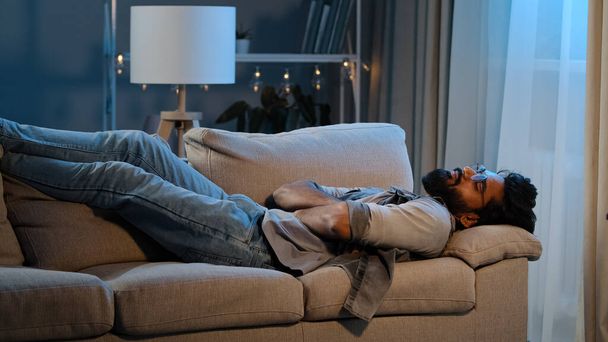Exhausted overloaded arabian man indian bearded guy unmotivated tired male came home after work flopped down on couch at night evening dark feels overworked sick ill person hard day no lack of energy - Фото, изображение