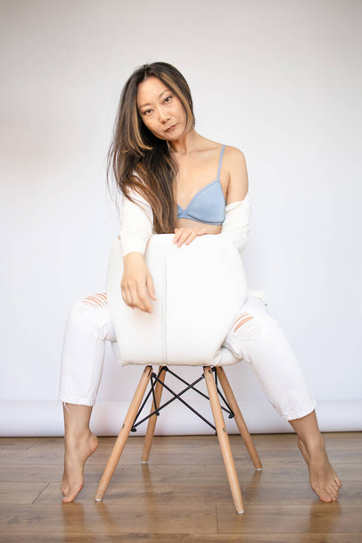 Portrait of asian woman wearing lingerie in studio. Young female model with black hair posing in fashion while sitting on stool looking at the camera. - Photo, Image
