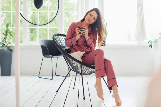 Cheerful young woman in pajamas sitting on the chair indoors at daytime. - Photo, Image
