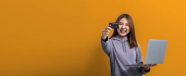 Portrét krásné asijské ženy držící kreditní kartu a notebook na izolovaném pozadí, portrét koncept použitý pro reklamu a nápisy, izolované přes žluté pozadí, kopírovací prostor. - Fotografie, Obrázek
