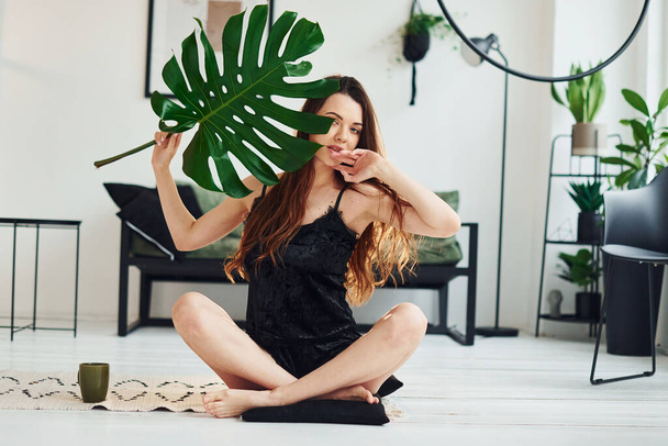 Young woman in pajamas sitting on the floor indoors at daytime and holding big green leaf. - Fotoğraf, Görsel