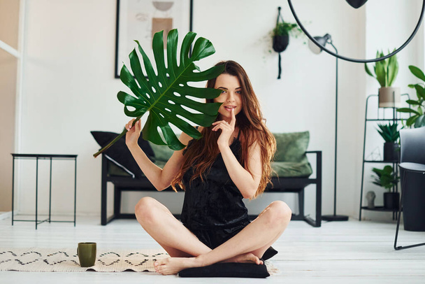 Young woman in pajamas sitting on the floor indoors at daytime and holding big green leaf. - Фото, изображение