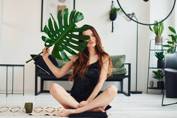 Young woman in pajamas sitting on the floor indoors at daytime and holding big green leaf. - Photo, image