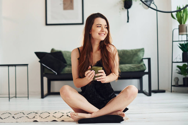 Young woman in pajamas sitting on the floor indoors at daytime with cup of drink. - Photo, image