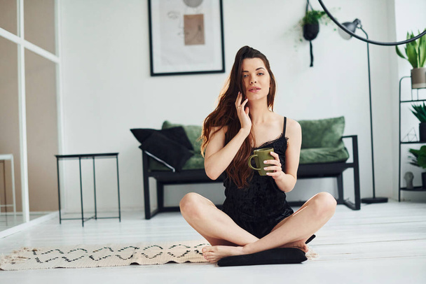 Young woman in pajamas sitting on the floor indoors at daytime with cup of drink. - Photo, image