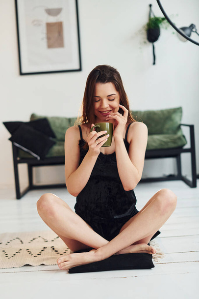 Young woman in pajamas sitting on the floor indoors at daytime with cup of drink. - 写真・画像