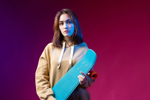 Young beautiful fashionable hipster girl dressed in a hoodie holds a skateboard in her hands on a purple studio background. - 写真・画像
