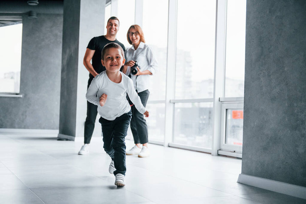 Woman in formal clothes and with camera in hand standing inside of empty room with man and little boy that running and having fun. - Foto, imagen