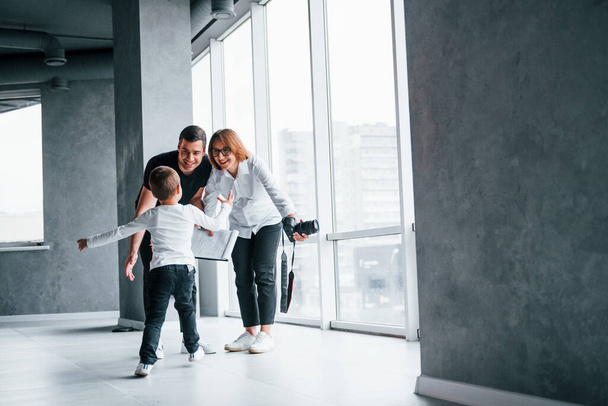 Woman in formal clothes and with camera in hand standing inside of empty room with man and little boy that running and having fun. - Фото, зображення