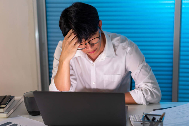 Concepto de exceso de trabajo El hombre sentado en su escritorio, quitándose las gafas y sosteniendo la cabeza mientras trabaja. - Foto, imagen