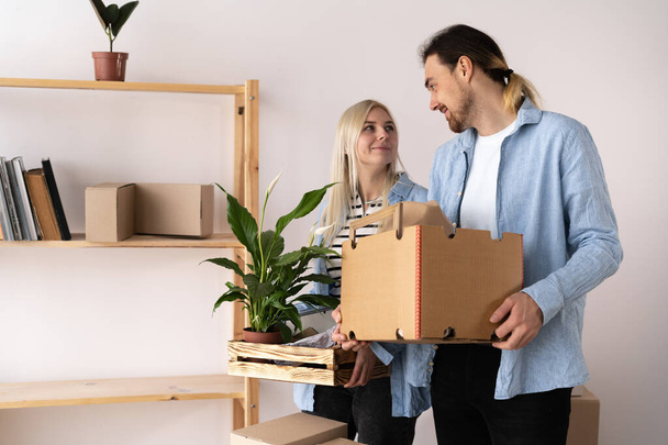 счастливая молодая пара, несущая картонные коробки и растения во время переезда в новую квартиру, мужчина и женщина переносят коробки в новый дом в день переезда, копировальное место - Фото, изображение