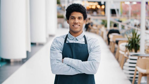 Jonge Afro-Amerikaanse man man restaurant cafe mannelijke werknemer op zoek naar camera poseren kruising armen ober verkoper dragen schort staan op het werk ruimte glimlachend vriendelijk. Beroep van kleine ondernemingen - Foto, afbeelding