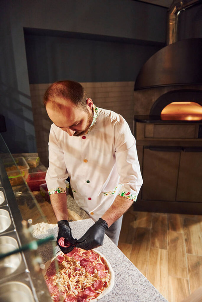 Šéfkuchař v bílém oblečení připravuje lahodnou pizzu na kuchyni restaurace. - Fotografie, Obrázek