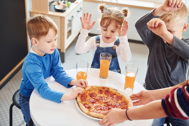 Három gyerek ül bent az asztalnál és együtt esznek pizzát narancslével.. - Fotó, kép