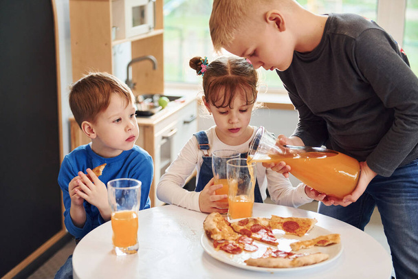 Three children sitting indoors by the table and eating pizza with orange juice together. - Foto, Imagem