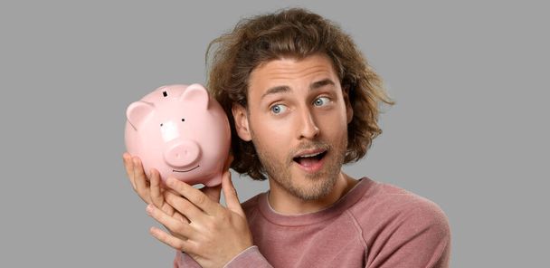 Curious man with piggy bank on grey background - Фото, изображение