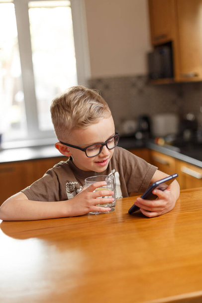 A five-year-old boy is in the kitchen, drinking water with lemon and looking at the phone. Caucasian boy in glasses drinking water from a glass in the kitchen - Foto, Imagem