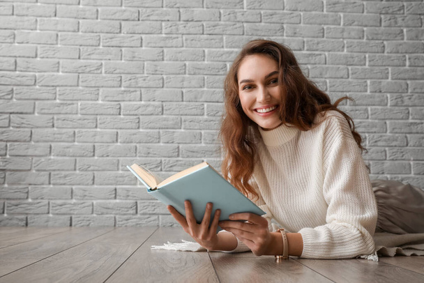 Beautiful young woman reading book on wooden floor near grey brick wall - Photo, Image