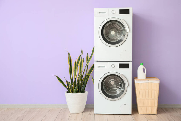 Secado moderno, lavadoras, cesta y planta de interior cerca de la pared violeta en la habitación - Foto, Imagen
