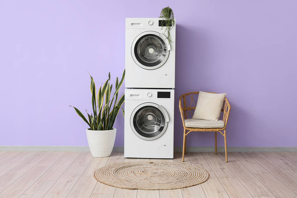 Asciugatura moderna, lavatrici, sedia di vimini e piante d'appartamento vicino alla parete viola all'interno della stanza - Foto, immagini