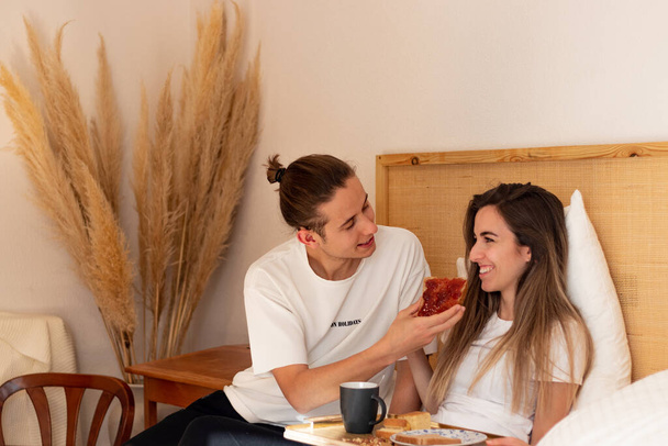 Junger Mann gibt seiner Frau im Bett einen Toast mit Erdbeermarmelade, während er im Zimmer frühstückt. Niedliches Liebespaar - Foto, Bild