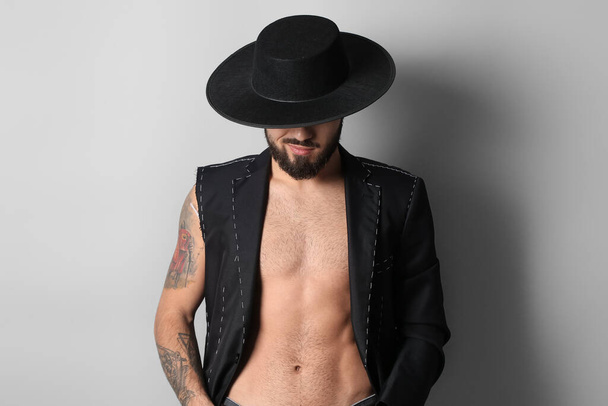 Young bearded man in half-finished jacket and stylish hat on light background - Foto, imagen