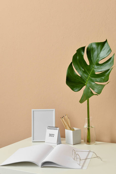 Elegante lugar de trabajo con marco de fotos en blanco, cuaderno y jarrón con hoja tropical cerca de la pared beige - Foto, imagen