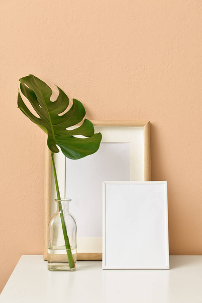 Stylish workplace with blank photo frames and vase with tropical leaf near beige wall - Foto, imagen