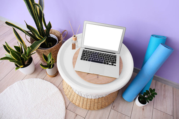 Tisch mit Laptop, Zimmerpflanzen und Yogamatten neben fliederfarbener Wand - Foto, Bild