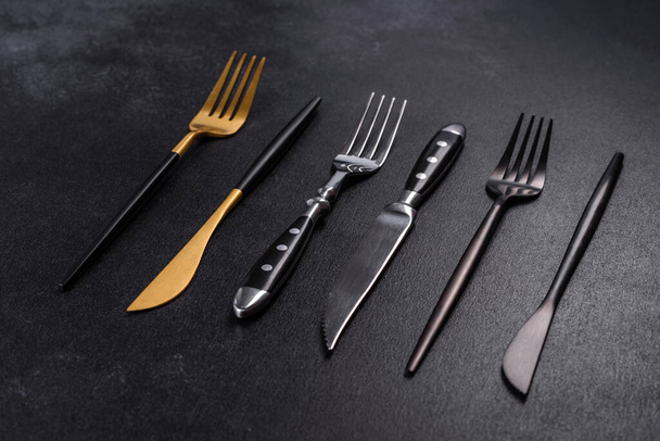 Diversi coltelli e forchette in oro nero e argento su fondo cemento scuro. Preparare il tavolo per il pranzo - Foto, immagini