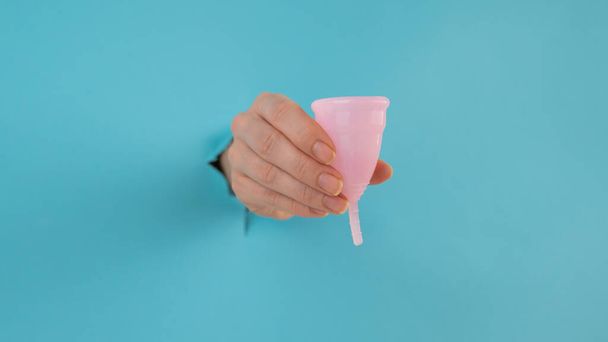A female hand sticking out of a hole from a blue background holds a pink menstrual cup.  - Foto, imagen
