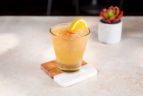 A view of a yellow orange peach colored alcoholic beverage, garnished with an orange slice and cherry, on ice.  - Photo, image