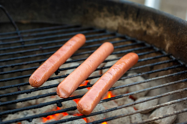 A view of three hot dogs on the grill. Glowing charcoals are underneath. - Foto, Imagem