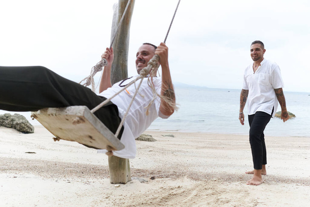 Гей, качающийся на пляжных качелях рядом со своей парой - Фото, изображение