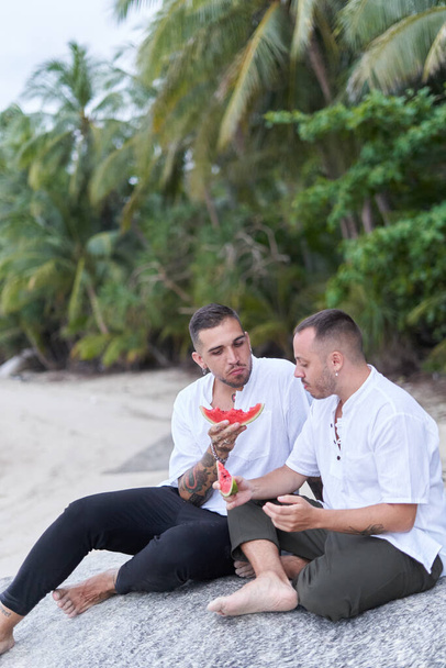 Κάθετη φωτογραφία ενός γκέι ζευγαριού που τρώει καρπούζι στην παραλία - Φωτογραφία, εικόνα